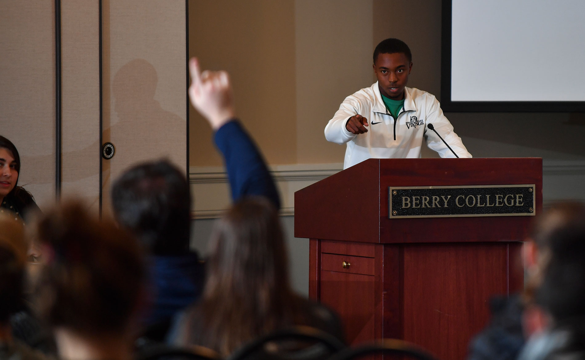 professor behind podium pointing to a student raising their hand in a crowd